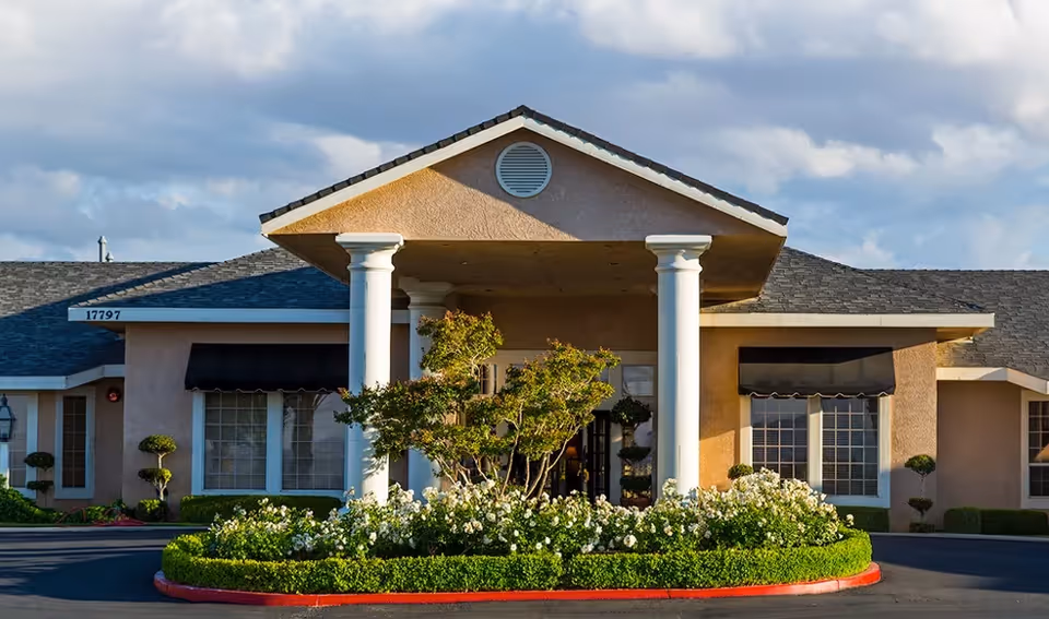 Front entrance of a single-story building with a columned portico, awninged windows, and manicured shrubs and flower beds.