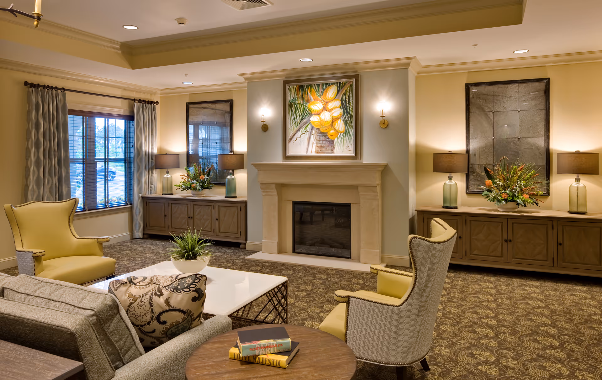 A cozy living room with a beige carpet and neutral walls featuring a fireplace in the center with a painting of yellow fruit above it. There are two yellow armchairs and a gray sofa with patterned cushions around a white coffee table. Two wooden sideboards with lamps, mirrors, and flower arrangements flank the fireplace. A window with patterned curtains is on the left side of the room.