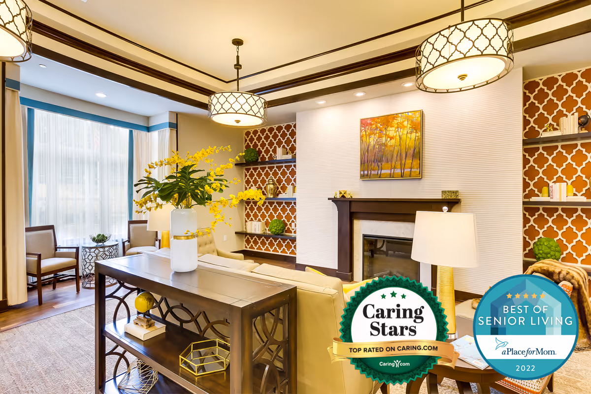 A bright and elegantly decorated living room with a beige sofa, a wooden console table with a vase of yellow flowers, two armchairs near a large window with sheer curtains, patterned wallpaper on the walls, built-in shelves with decorative items, a fireplace with a colorful painting above it, and two hanging ceiling lights.