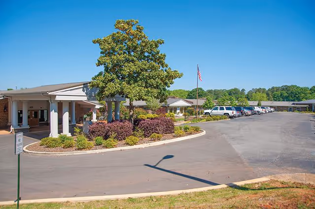 Exterior view of Jacksonville Health and Rehabilitation, LLC showing a single-story building with a covered entrance, landscaped greenery including a tree and bushes, an American flag on a flagpole, and a parking lot with several parked cars under a clear blue sky.