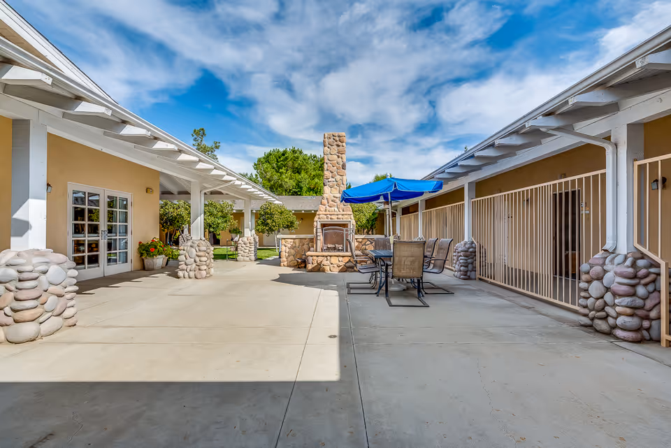 Outdoor courtyard area of a senior living facility with a stone fireplace in the center, surrounded by patio tables and chairs with a blue umbrella. The courtyard is flanked by buildings with beige walls, white trim, and stone pillars. The sky is partly cloudy with blue patches.