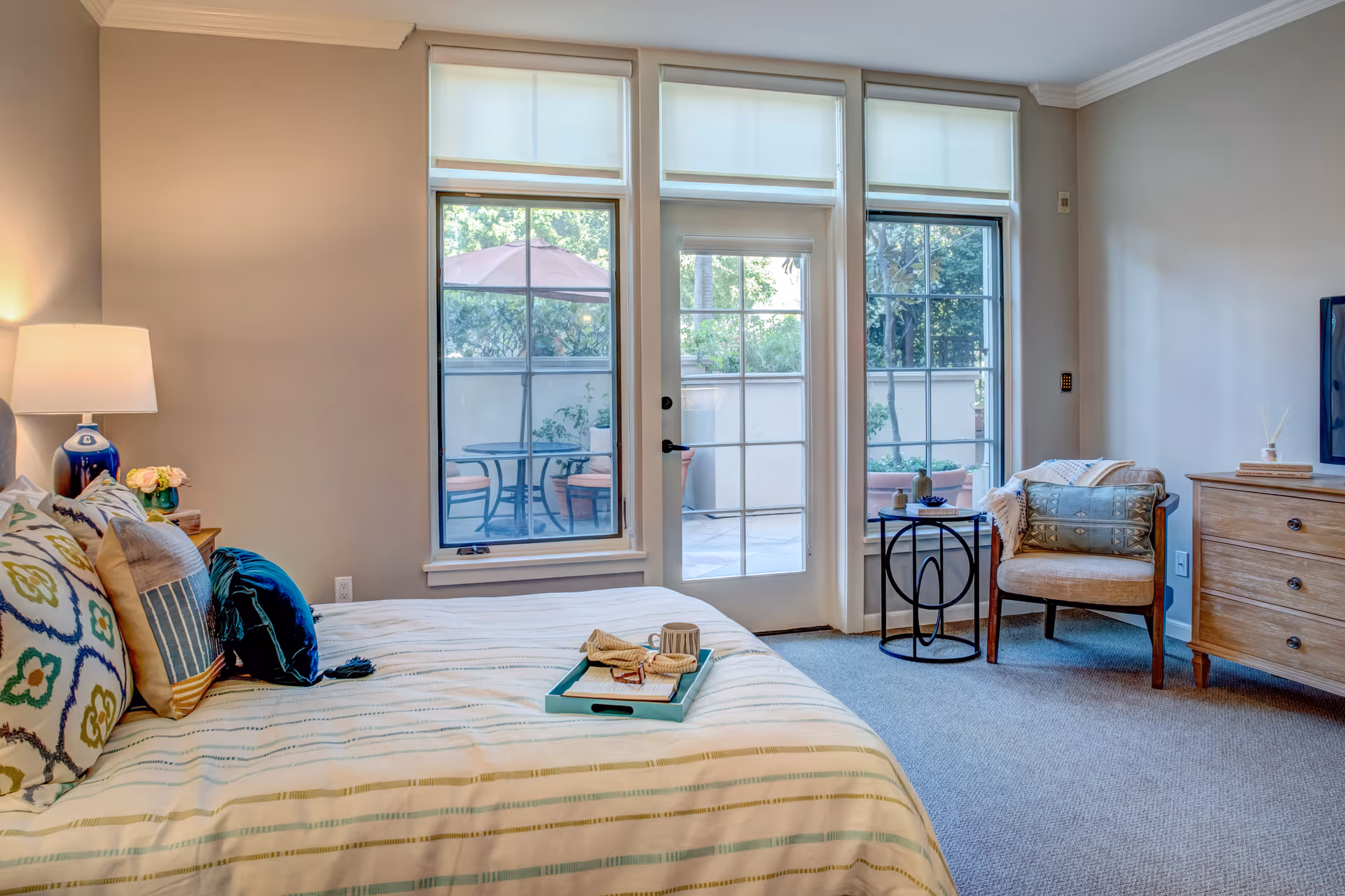 Bright furnished bedroom with a bed, armchair, dresser, and glass doors opening to a small patio.