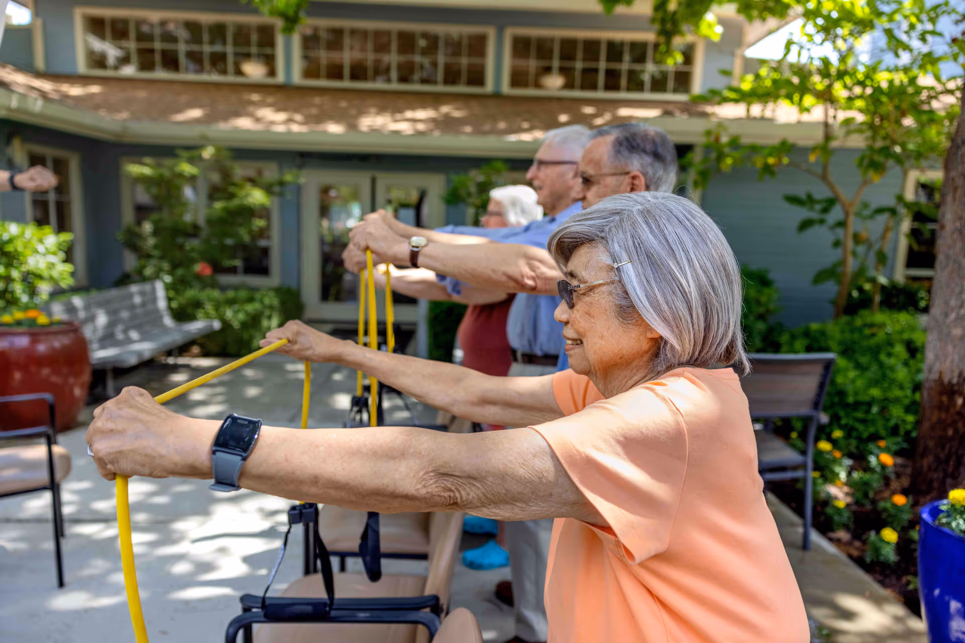 A group of elderly people exercising outdoors using yellow resistance bands, standing in a row with their arms extended forward. They are in a garden area with greenery and a building in the background.