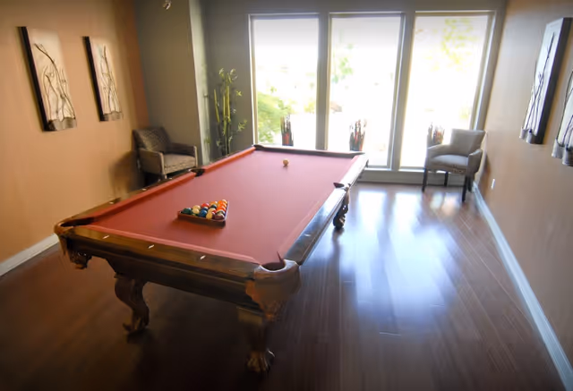 A room with a red felt pool table centered on a wooden floor, two armchairs placed near large windows letting in natural light, and decorative wall art on the walls.