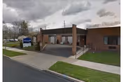 Exterior view of Kingston Rehabilitation & Nursing Center - Bedrock Care showing a brick building with a covered entrance and a sign on the lawn near the sidewalk. The sky is cloudy and there is a paved road in front of the building.
