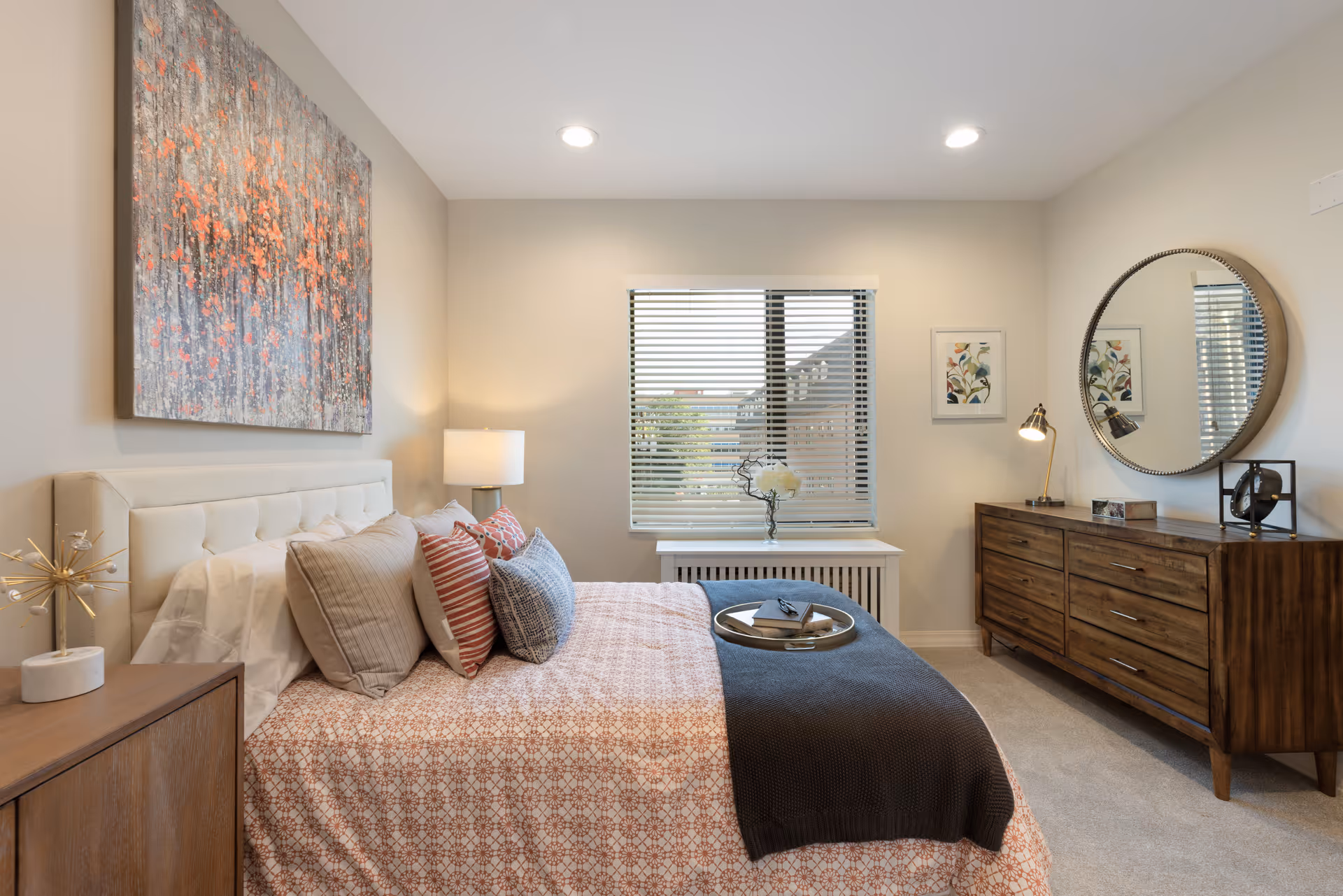 Well-lit furnished bedroom with a bed, bedside table and lamp, a dresser with round mirror, and a window with blinds.