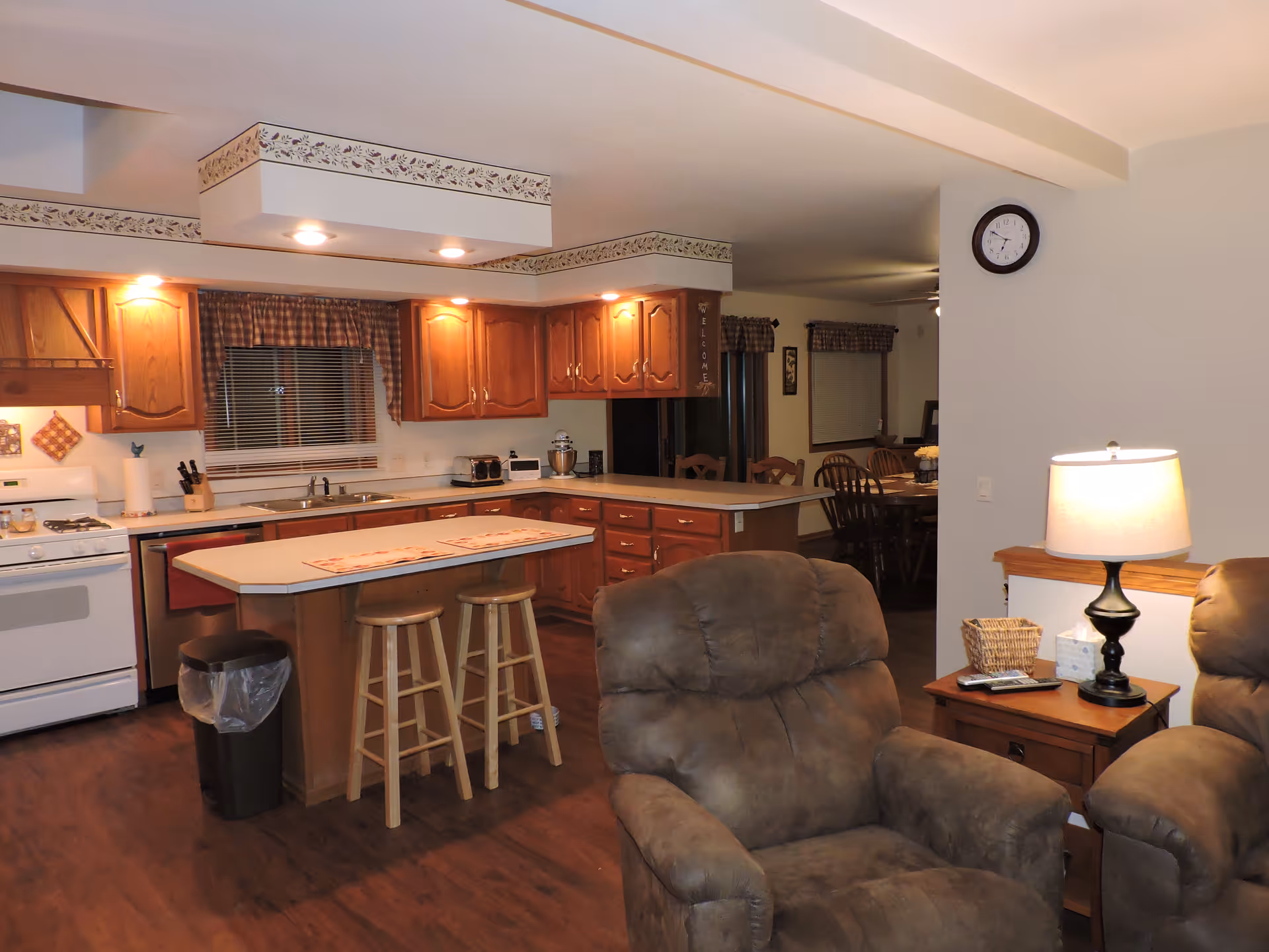 Interior view of a senior living facility showing a cozy kitchen with wooden cabinets, a white stove, a kitchen island with two wooden stools, and a living area with two cushioned armchairs and a side table with a lamp and tissue box. In the background, a dining area with a wooden dining table and chairs is visible.