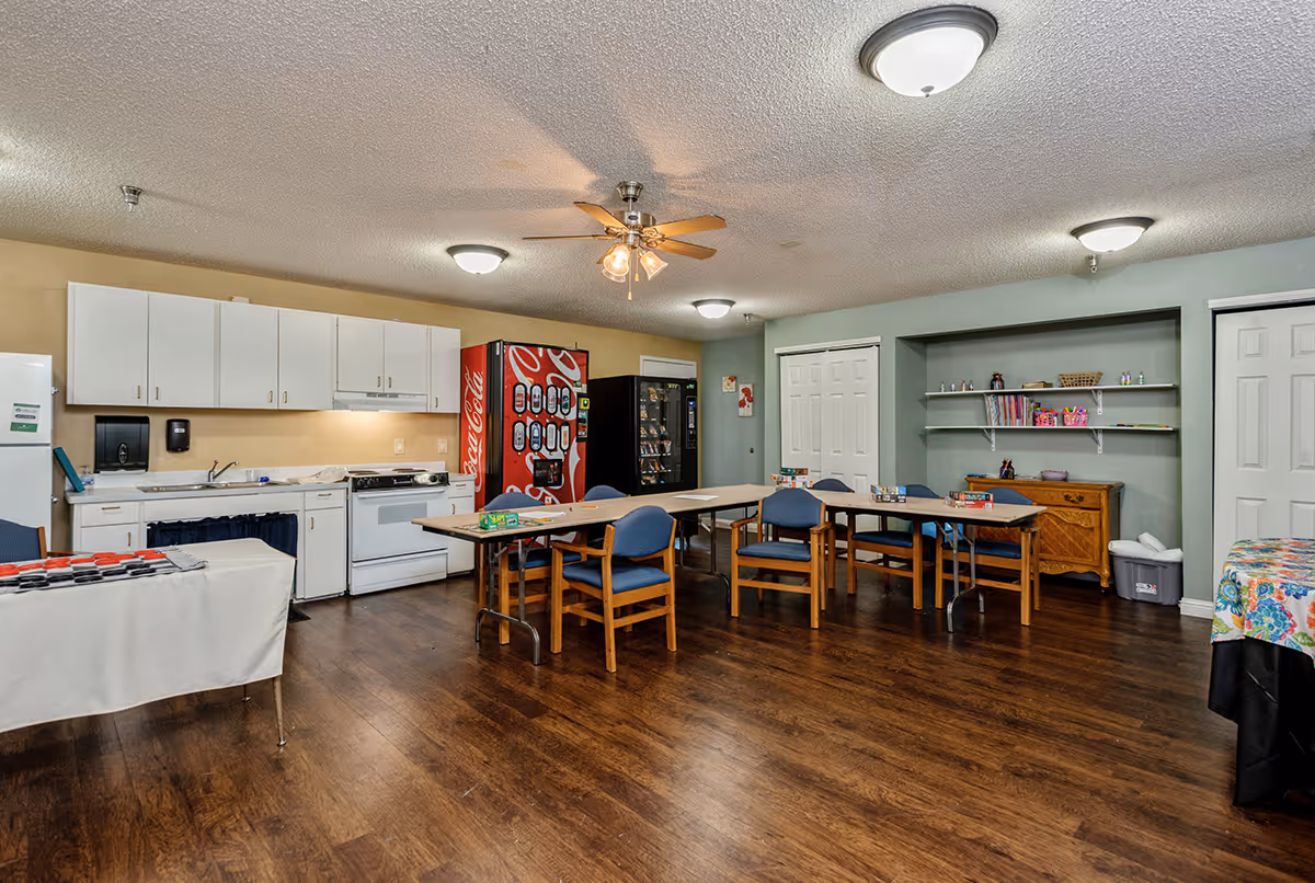 A communal room with a kitchenette area featuring white cabinets, a stove, and a refrigerator. There are two vending machines, several tables with blue chairs arranged around them, and shelves with books and small items. The room has wooden flooring, ceiling lights, and a ceiling fan with lights.