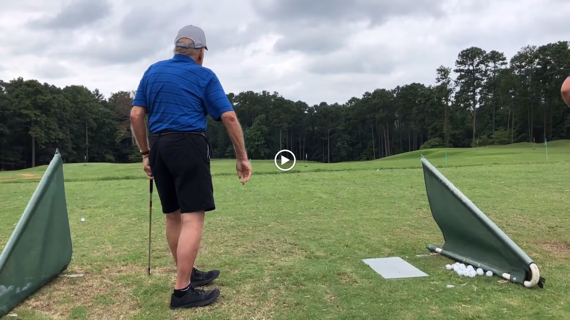 An elderly man wearing a blue shirt, black shorts, and a gray cap is standing on a grassy golf practice range holding a golf club. There are golf balls scattered on the ground near two green barriers, and trees line the background under a cloudy sky.