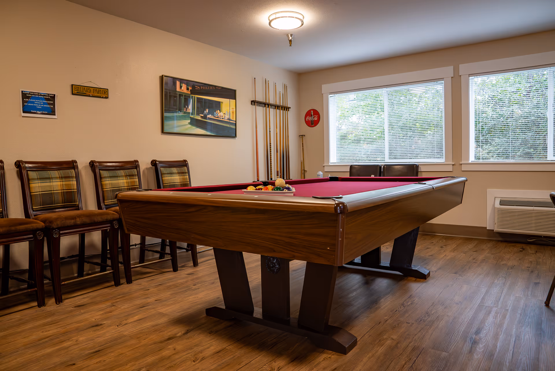 A billiard parlor room with a pool table featuring a red felt surface and a rack of billiard balls set up for a game. There are several wooden chairs with plaid cushions lined up against the wall, a rack holding pool cues, a framed painting on the wall, and two large windows with blinds letting in natural light. The room has wooden flooring and a ceiling light fixture.