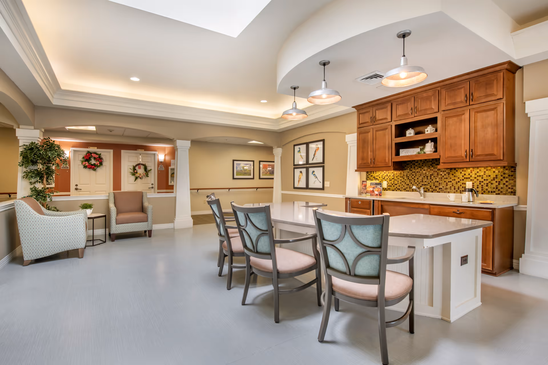 A bright and spacious common area in a senior living facility featuring a kitchen with wooden cabinets, a mosaic tile backsplash, and a large island with four chairs. The room has a light gray floor, recessed lighting, and three pendant lights above the island. In the background, there are two armchairs with a small table between them, a potted plant, and two doors decorated with wreaths. The walls are painted in neutral tones with framed bird artwork.