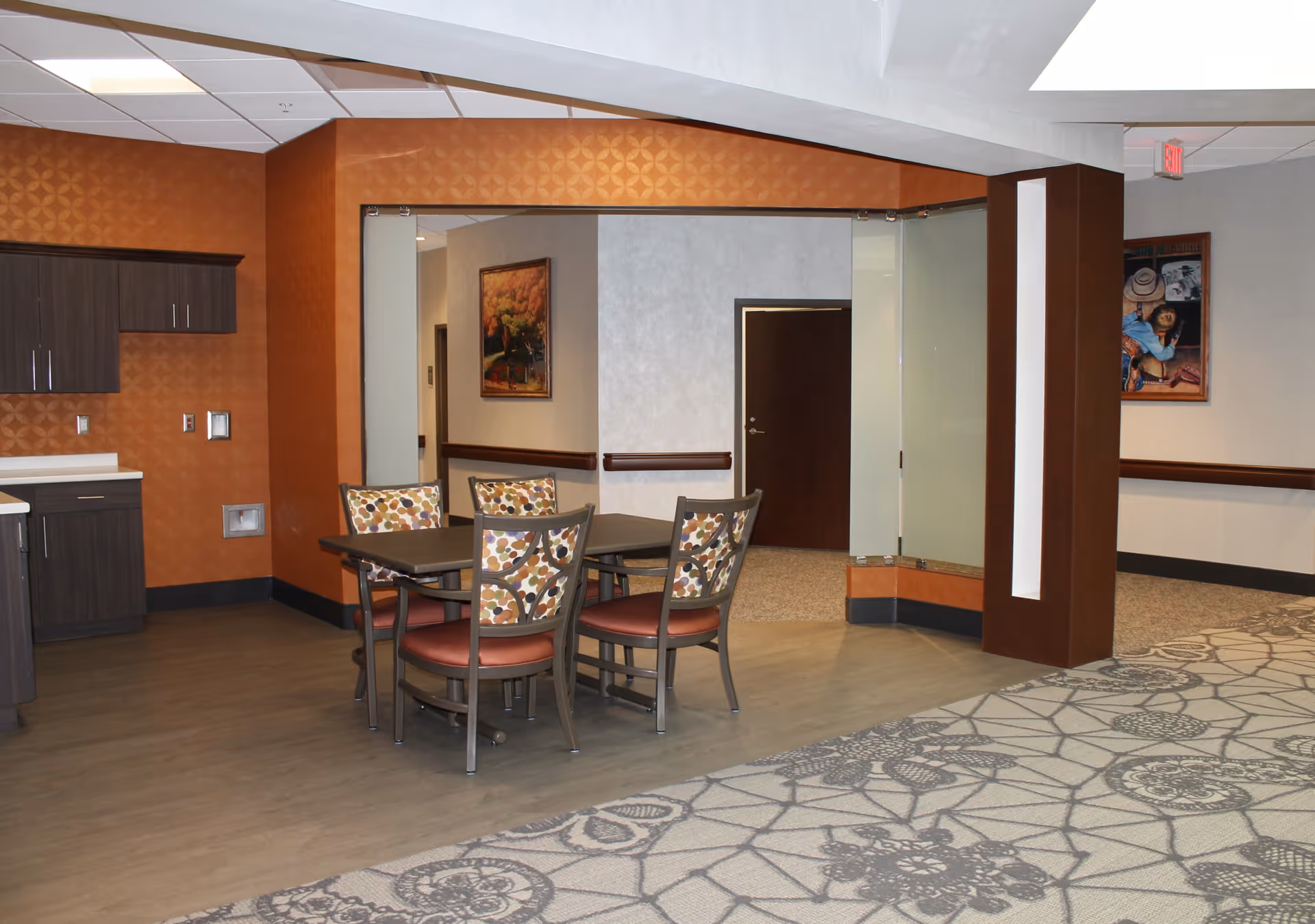 Interior view of a common area in a rehabilitation center featuring a small dining table with four chairs that have patterned cushions. The room has a mix of wood and carpet flooring, orange and beige walls, and some framed artwork. There are cabinets and a countertop on the left side.