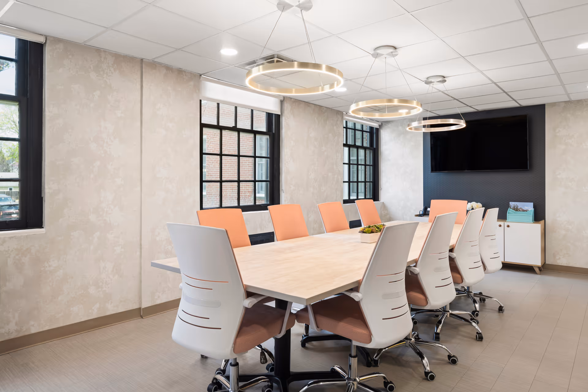 Bright conference room with a long light wood table surrounded by white rolling chairs with peach cushions, circular pendant lights, windows, and a wall-mounted TV.