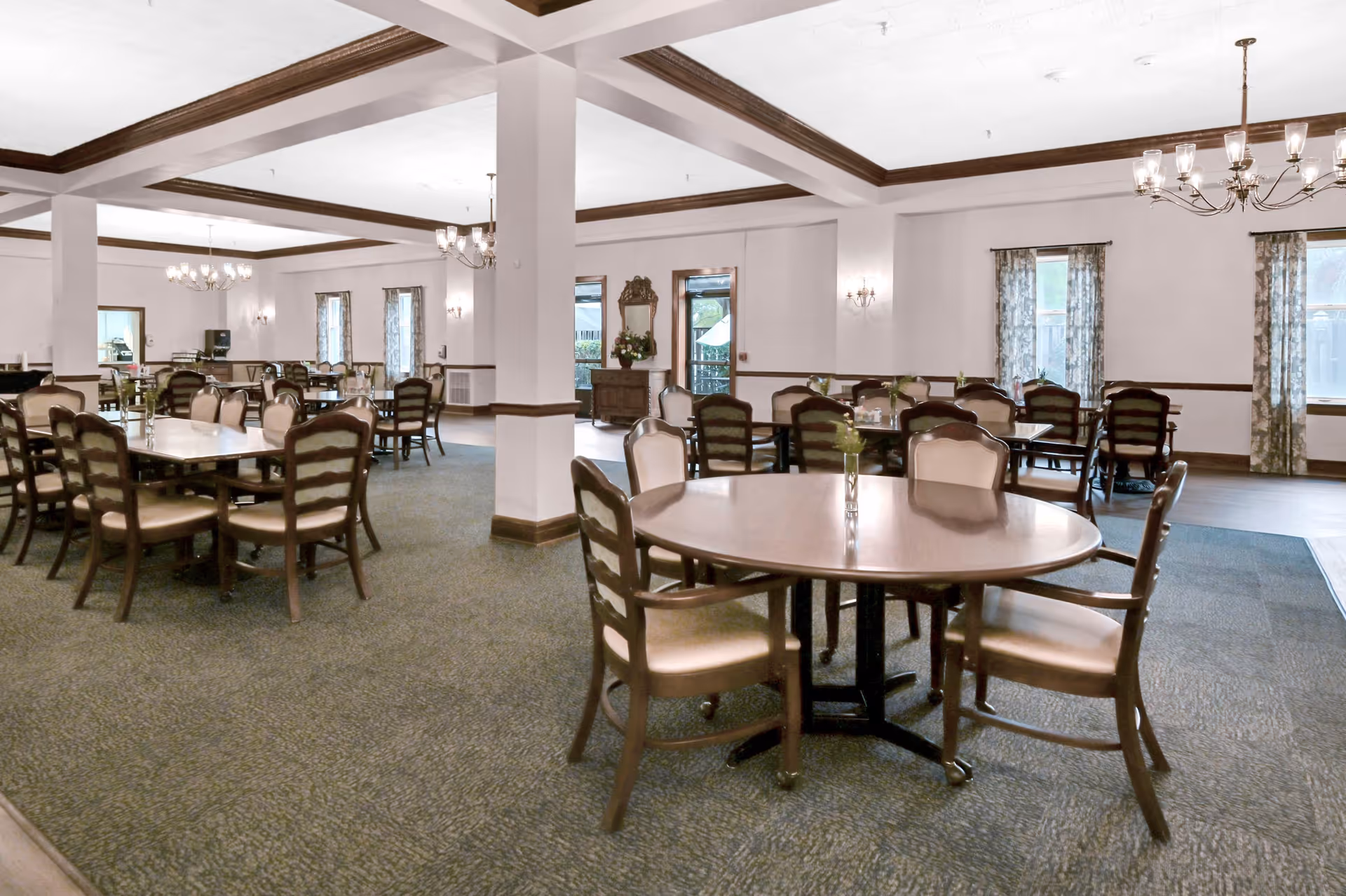 A spacious dining room with multiple wooden tables and chairs arranged neatly. The room features carpeted flooring, white walls with wooden trim, several windows with patterned curtains, and chandeliers hanging from a coffered ceiling providing warm lighting.