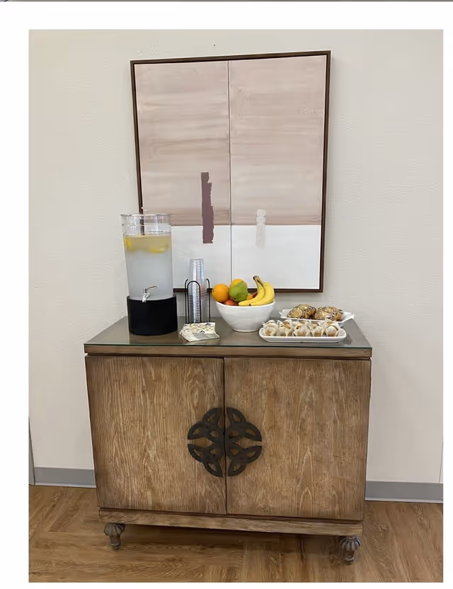 Wooden side cabinet topped with a lemon water dispenser, a bowl of fruit and pastries beneath abstract wall art.