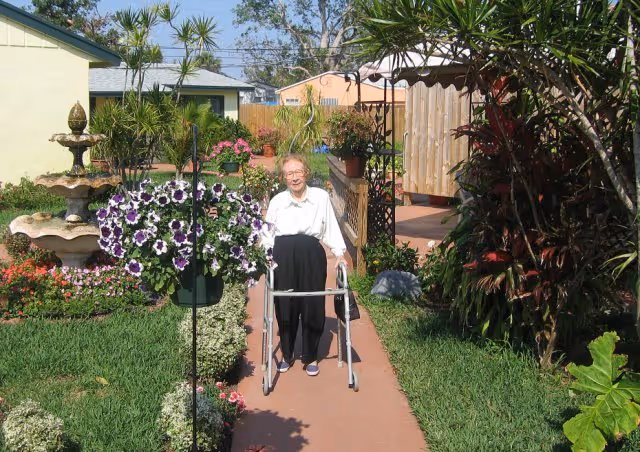 An elderly person using a walker is walking on a paved path through a garden area with green grass, flowering plants, and a multi-tiered water fountain on the left side. There are houses and trees in the background under a clear sky.