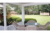Covered outdoor patio area with a round glass table and four wicker chairs, overlooking a well-maintained grassy lawn with trees and small buildings in the background.