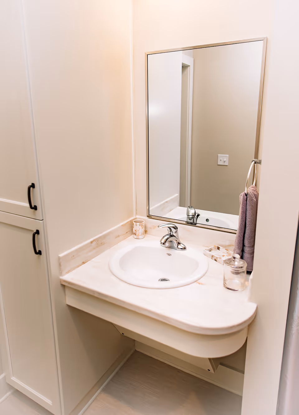 A clean bathroom sink area with a white countertop and a silver faucet. There is a large rectangular mirror above the sink, a purple hand towel hanging on a ring to the right, a small decorative cup, a soap dish, and a glass jar on the countertop. To the left, there is a white cabinet with black handles.