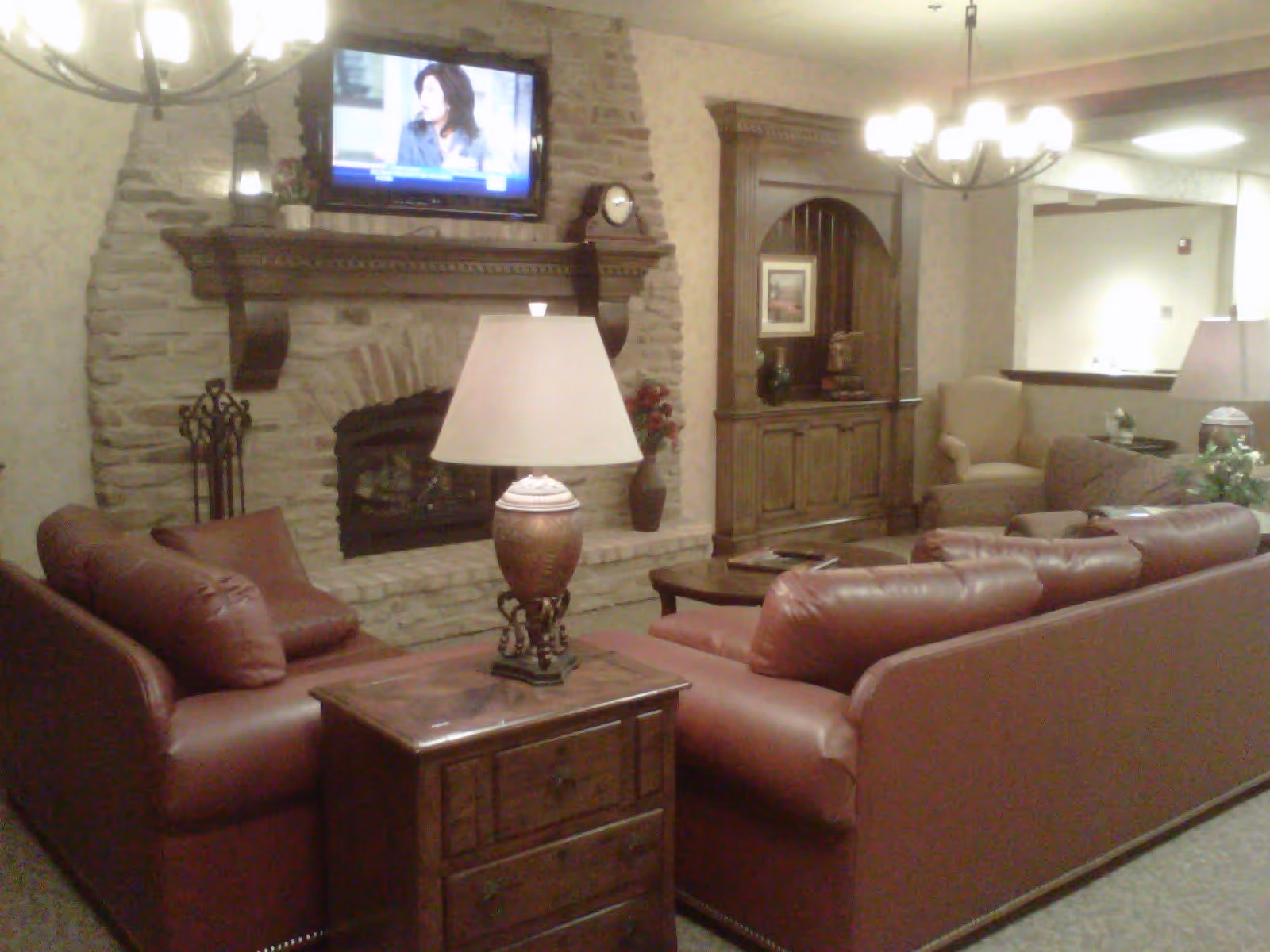 A cozy living room area with two red leather sofas facing a stone fireplace with a mounted flat-screen TV above it. A wooden side table with a decorative lamp is placed between the sofas. There is a wooden cabinet with shelves and decorative items on the right side, and an armchair is visible in the background. The room is warmly lit with ceiling lights and a chandelier.