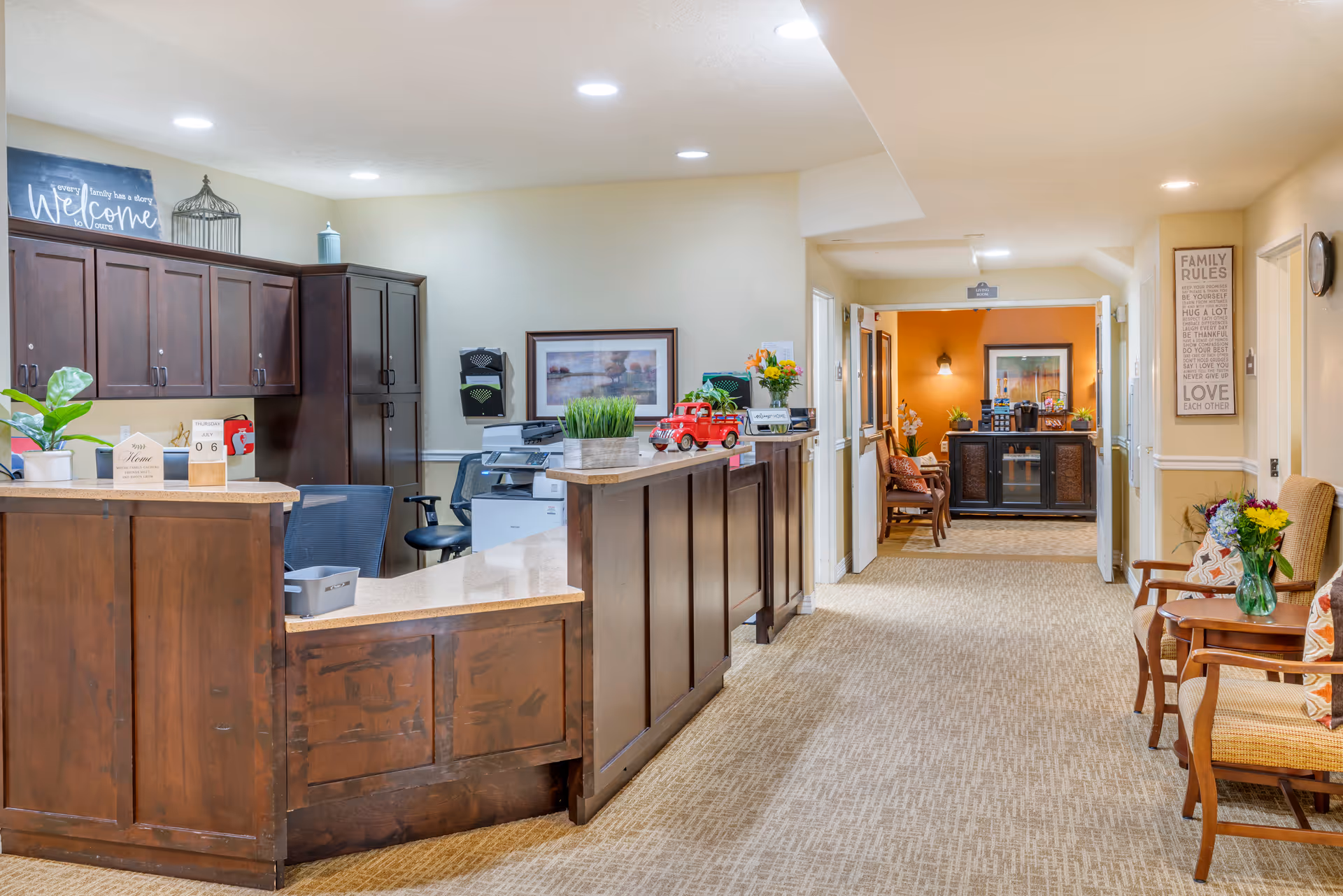 Reception area in a senior living facility with a wooden front desk, office chairs, and cabinets. The hallway extends to a seating area with chairs, a table with flowers, and a sign on the wall with family rules. The space is well-lit with ceiling lights and decorated with plants and framed artwork.