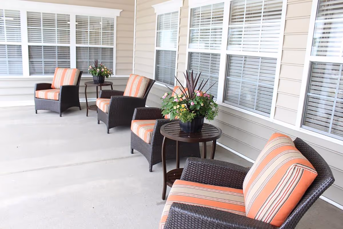 A covered outdoor patio area with four wicker armchairs featuring orange and beige striped cushions. Two small round wooden tables hold potted plants with green foliage and flowers. The patio is adjacent to a building with beige siding and multiple windows with white blinds.