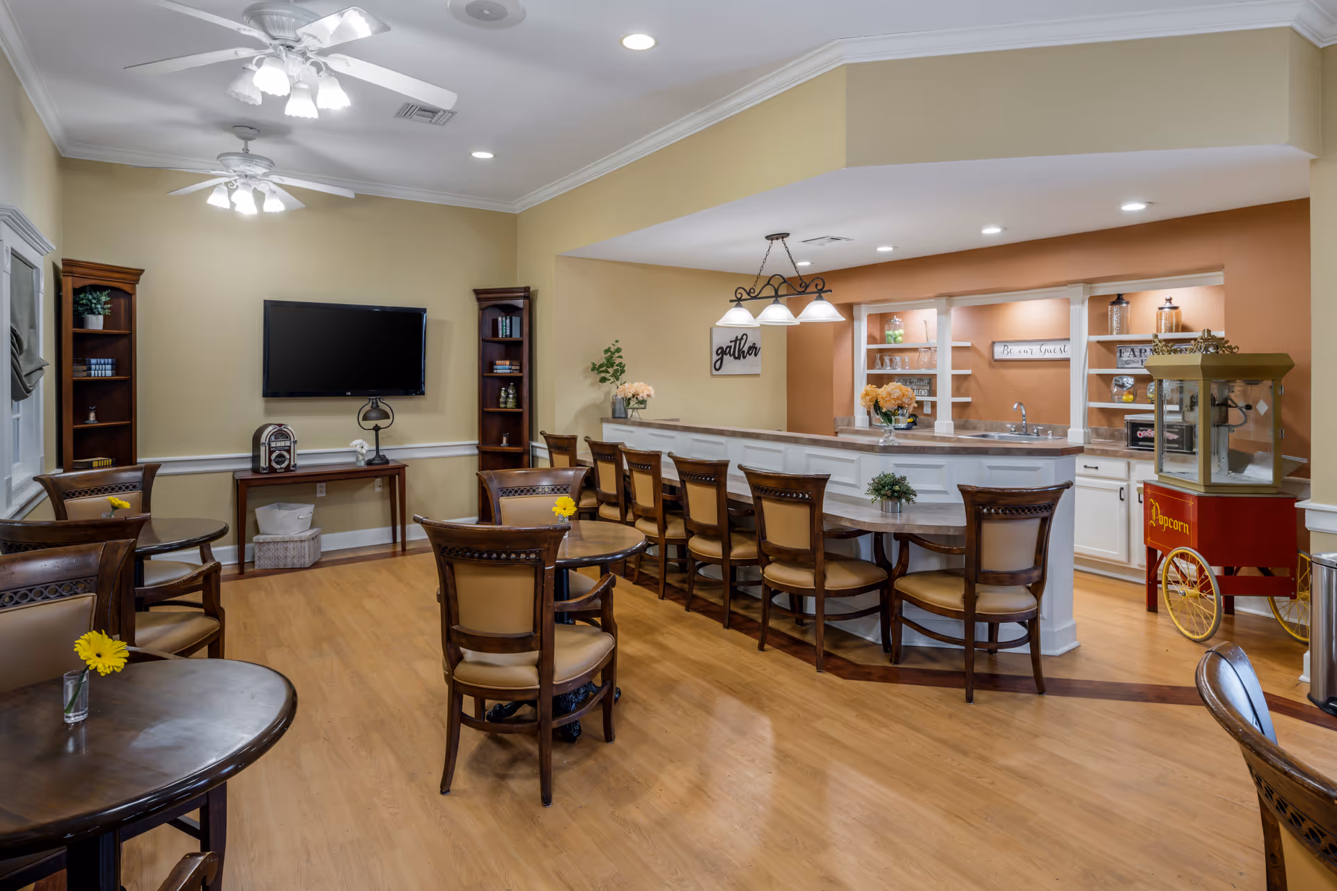 Communal dining and lounge area with a long bar, multiple round tables and chairs, a wall-mounted TV, and a popcorn cart.
