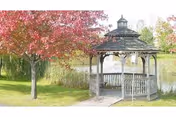 A wooden gazebo beside a pond surrounded by grass and trees with red autumn foliage.