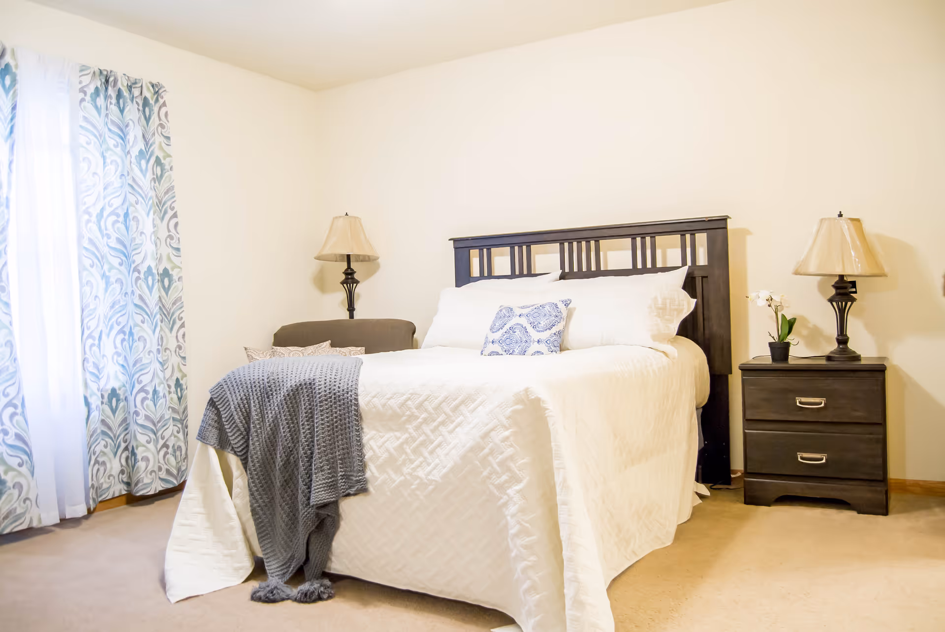 A bright bedroom with a neatly made bed featuring white bedding and a blue decorative pillow. There is a gray throw blanket draped over the foot of the bed. On the right side of the bed is a dark wooden nightstand with two drawers, a lamp, and a small potted plant. On the left side, there is a matching lamp and a cushioned chair. The room has light-colored walls and carpet, with patterned curtains covering a window on the left.