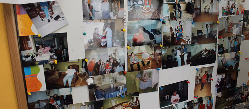 A bulletin board filled with numerous pinned photographs showing various people in different indoor settings, likely residents and staff of an assisted living facility engaged in social activities and gatherings.