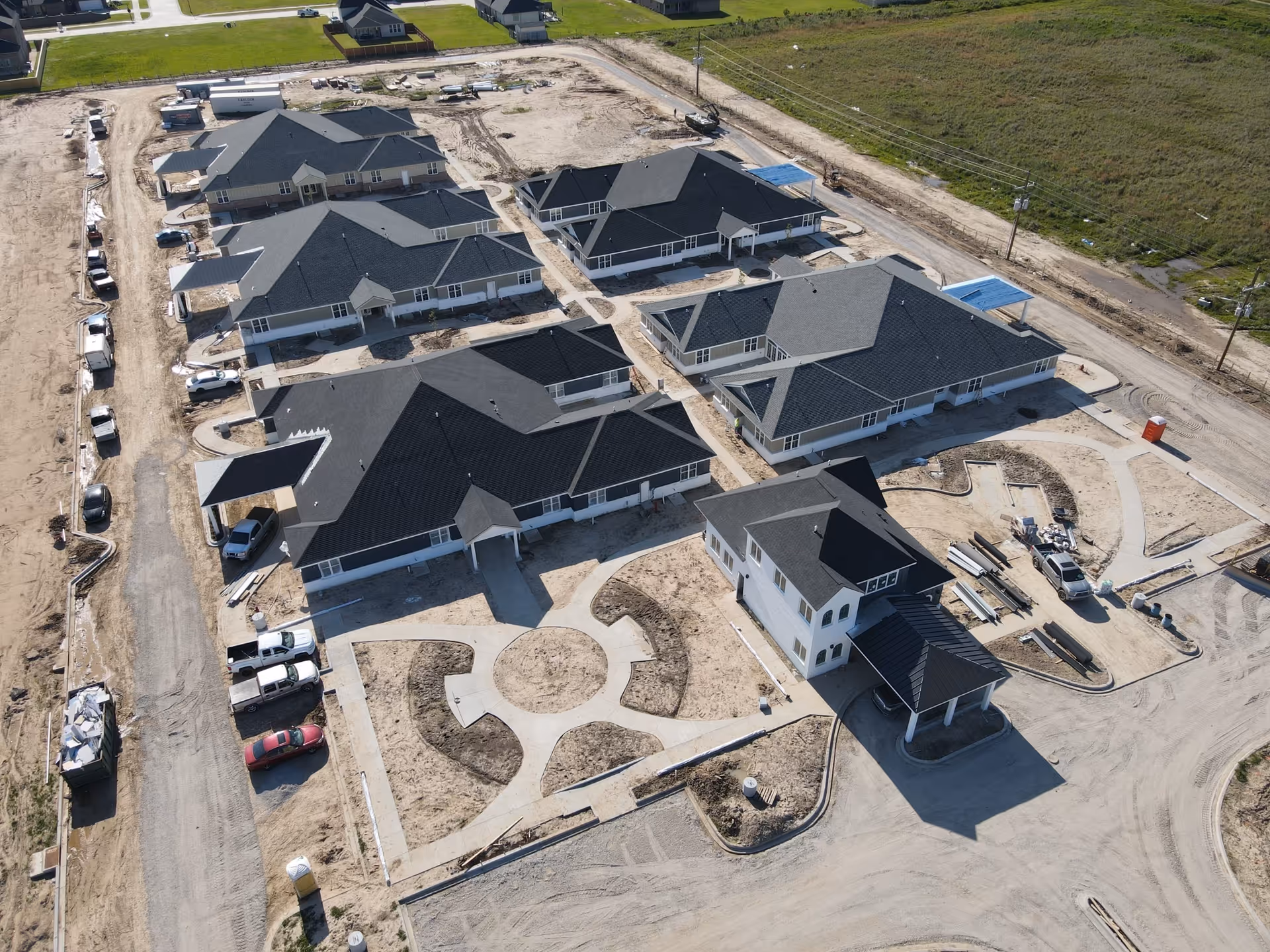 Aerial view of a senior living facility under construction with multiple single-story buildings with dark roofs arranged around paved walkways and open areas. Construction vehicles and materials are visible around the site, with some green grass and a few houses in the background.