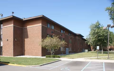 Brick senior housing building exterior with trees, sidewalk, and a parking lot.