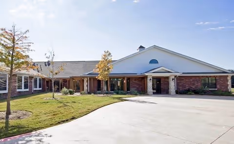 Exterior view of a single-story brick building with a white gabled roof and a covered entrance, surrounded by a grassy area with a few small trees under a clear blue sky.