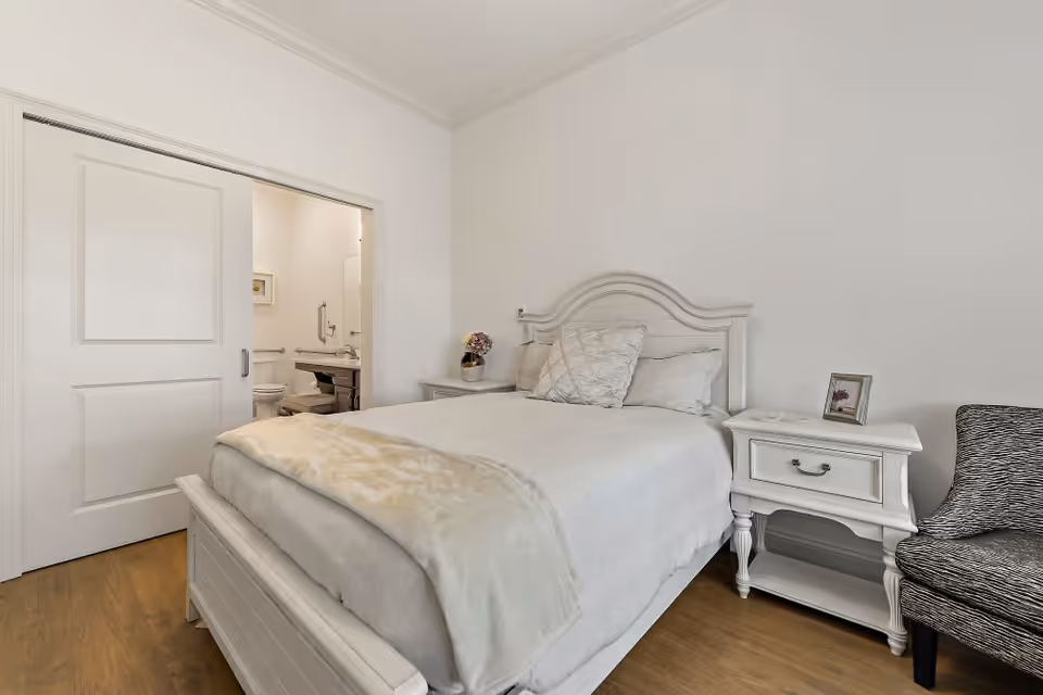 A cozy bedroom with a white wooden bed featuring a headboard and footboard, dressed in white bedding and a beige throw blanket. There are two white nightstands on either side of the bed, one with a small flower arrangement and the other with a framed photo. A patterned armchair is positioned next to one nightstand. A sliding door is partially open, revealing a bathroom with a toilet and sink.