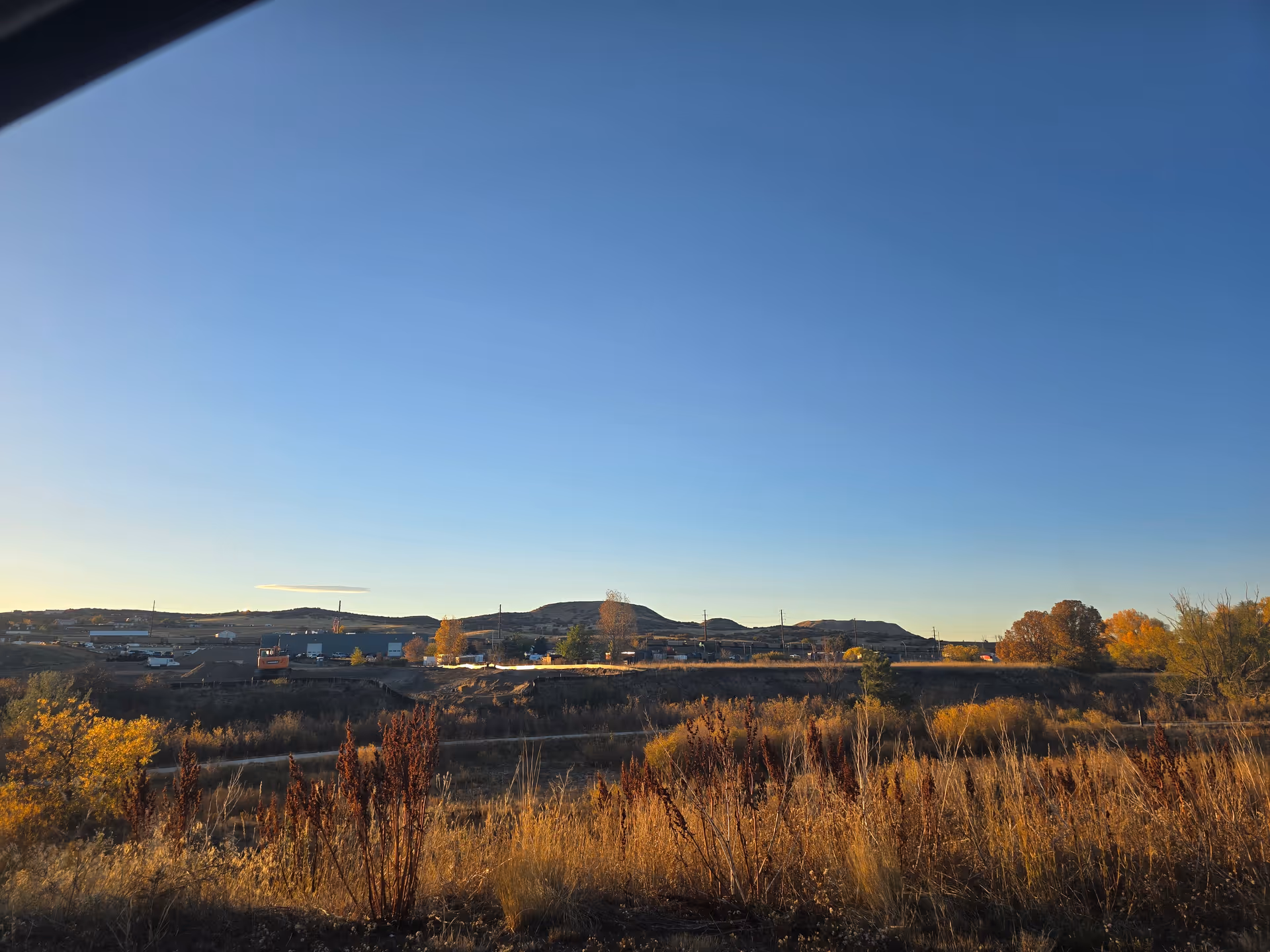 A landscape view showing dry grass and shrubs in the foreground with a clear blue sky above. In the distance, there are low hills and some buildings, possibly part of a small town or industrial area.