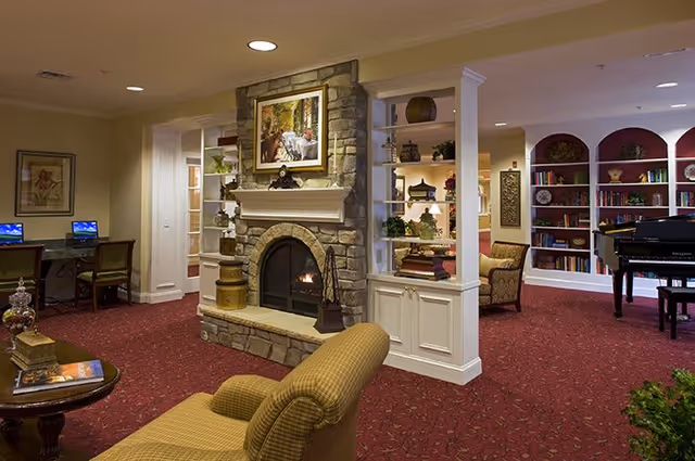 A cozy senior living facility common area with a stone fireplace in the center, flanked by built-in shelves with decorative items. There is a comfortable armchair in the foreground, a table with books and decor, a computer workstation with two chairs on the left, and a grand piano near a bookshelf on the right. The room has warm lighting and red patterned carpet.