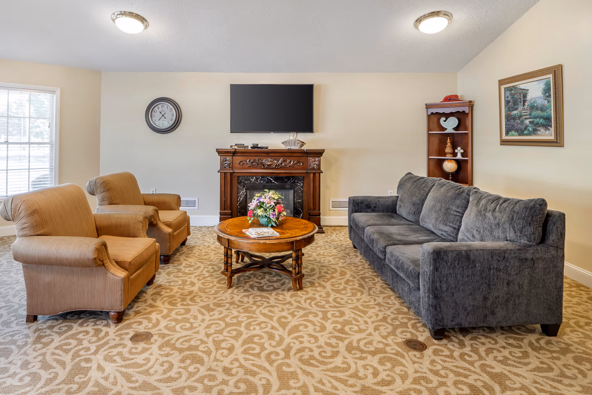 Cozy common living room with a gray sofa, two tan armchairs, a round wooden coffee table with flowers, and a wall-mounted TV above a decorative fireplace.