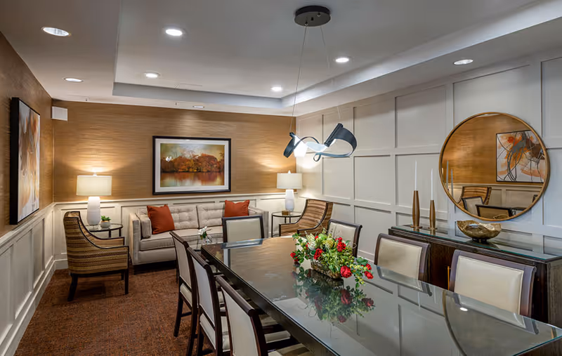 A stylish communal dining room with a long glass table surrounded by upholstered chairs, a seating area with a sofa and armchairs, artwork, and modern lighting.