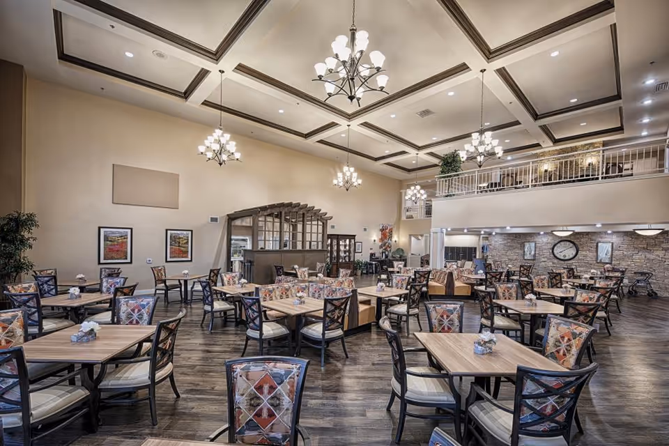 Spacious dining room with multiple wooden tables and chairs featuring patterned upholstery. The room has high ceilings with decorative beams and several chandeliers providing warm lighting. There is a mezzanine level with a railing overlooking the dining area, and the walls are decorated with framed artwork and a large clock. The floor is wooden, and the overall atmosphere is inviting and well-lit.