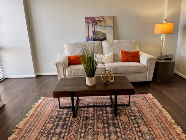 A cozy living room with a beige sofa adorned with two orange and one white cushion. In front of the sofa is a dark wooden coffee table with a potted plant and a decorative gold orb. The floor features a patterned area rug with fringe edges. A side table with a lamp and a small decorative item is positioned to the right of the sofa. A colorful painting is hung on the wall above the sofa.