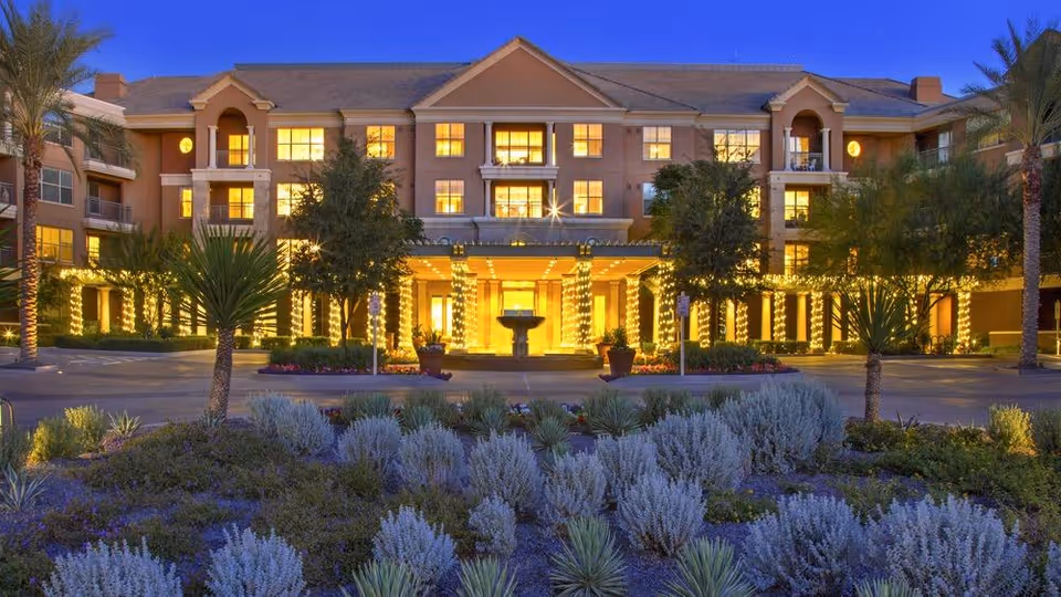 Well-lit, multi-story senior living building front at dusk with a decorated entrance, palm trees and landscaped shrubs in the foreground.