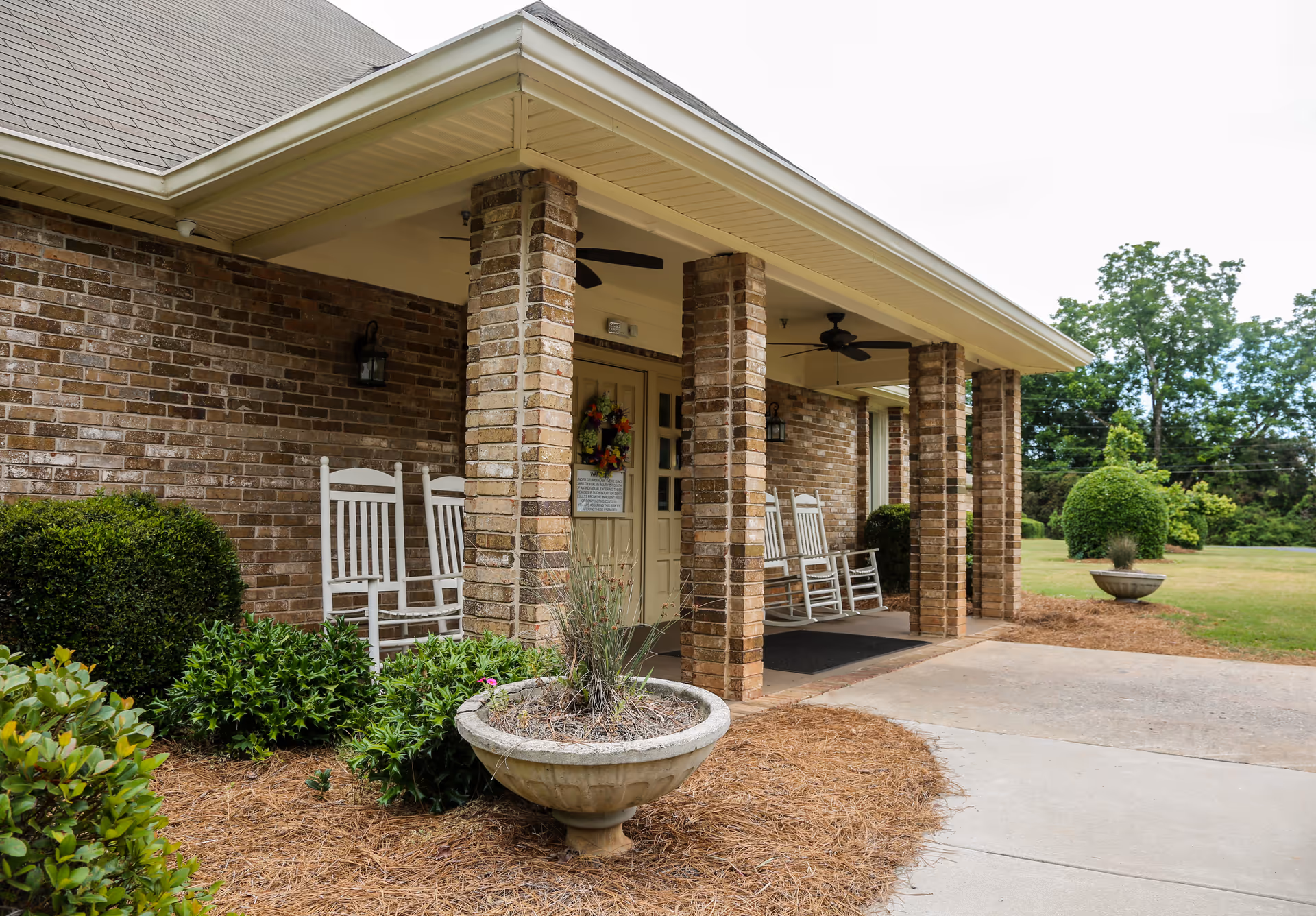 Front porch of a brick building with a covered entrance supported by brick columns. There are white rocking chairs on the porch, ceiling fans, and decorative plants in large planters. The surrounding area has neatly trimmed bushes and a grassy lawn with trees in the background.
