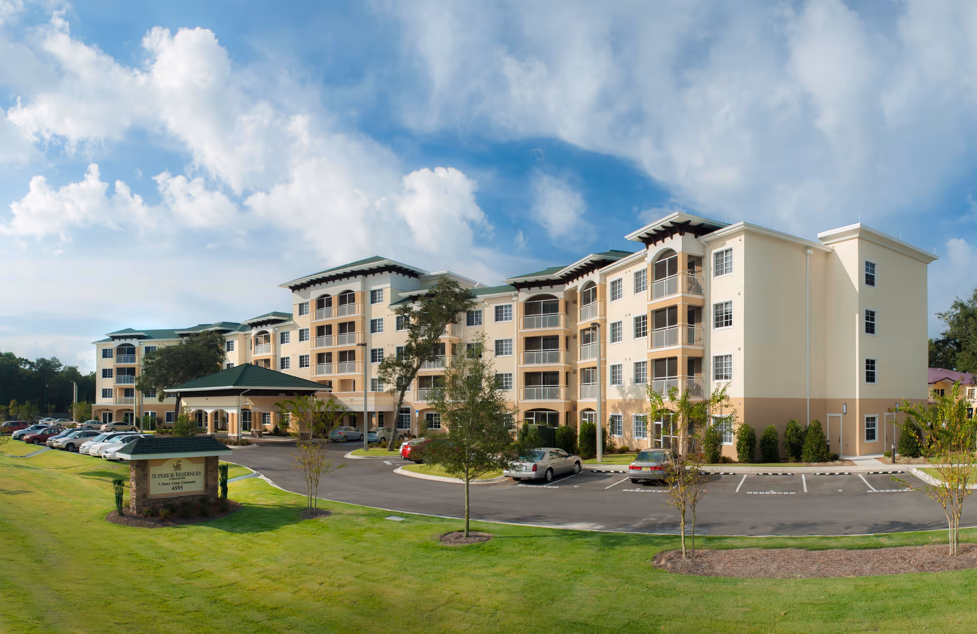 Exterior view of a large, multi-story senior living facility building with beige walls and green roofs. The building has multiple balconies and windows, surrounded by a well-maintained lawn and a parking lot with several cars. The sky is partly cloudy with blue patches.