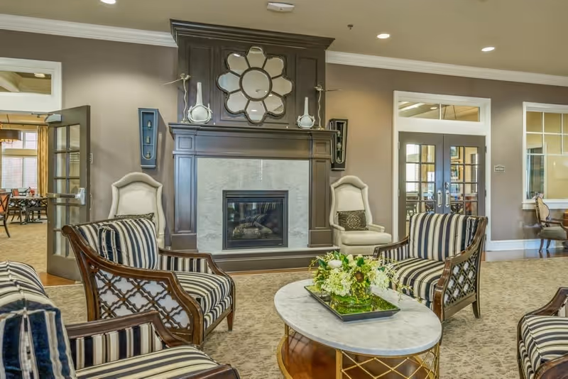 A cozy living room area featuring a fireplace with a decorative mirror above it, two high-back armchairs on either side, and several striped upholstered chairs arranged around a round marble coffee table with a floral centerpiece. The room has beige walls, carpeted flooring, and glass doors leading to other rooms.