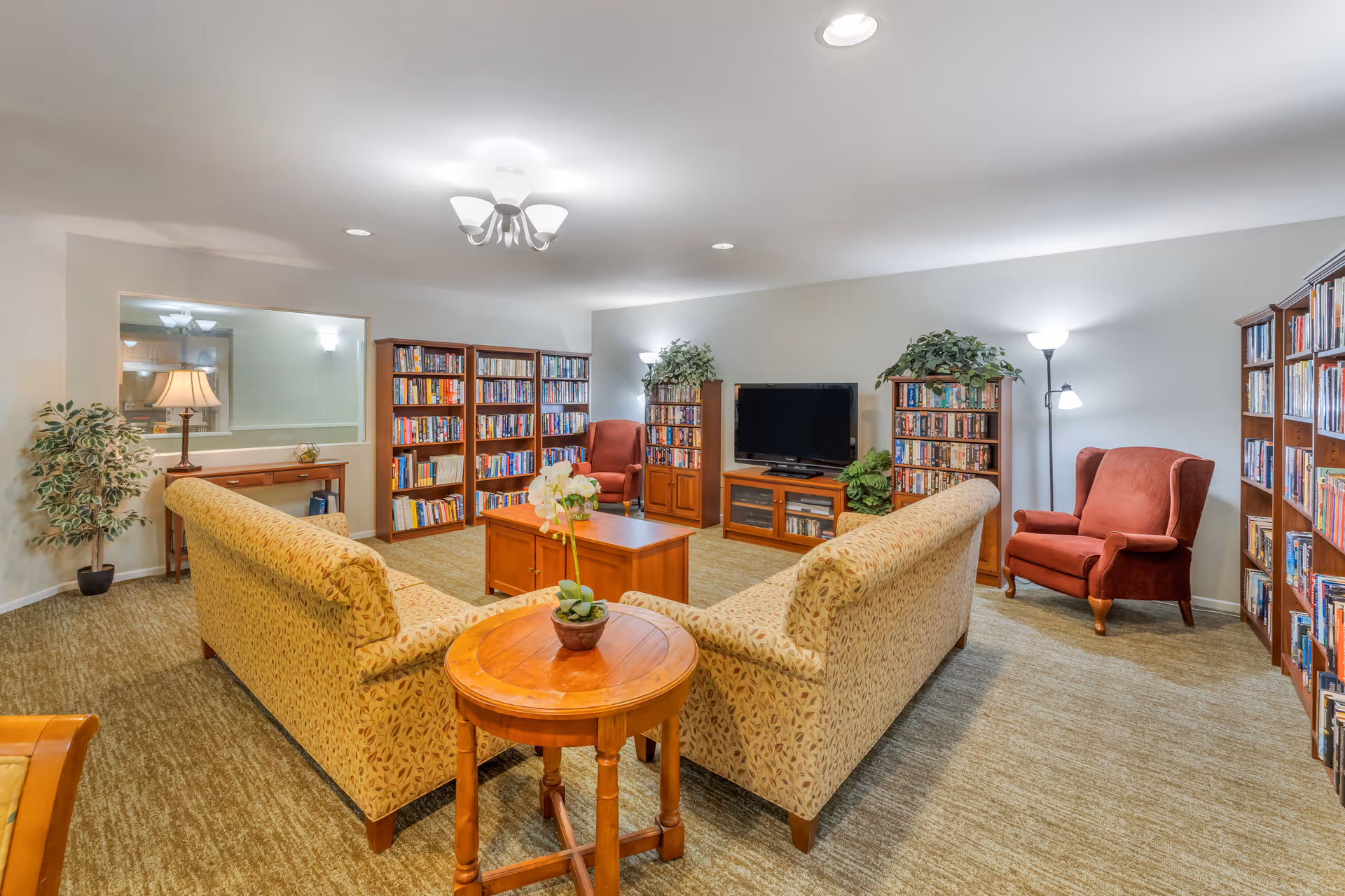 A cozy living room area with two patterned sofas arranged around a wooden coffee table. The room features multiple bookshelves filled with books, a flat-screen TV on a wooden stand, two red armchairs, and several potted plants. The lighting includes ceiling lights and floor lamps, creating a warm and inviting atmosphere.