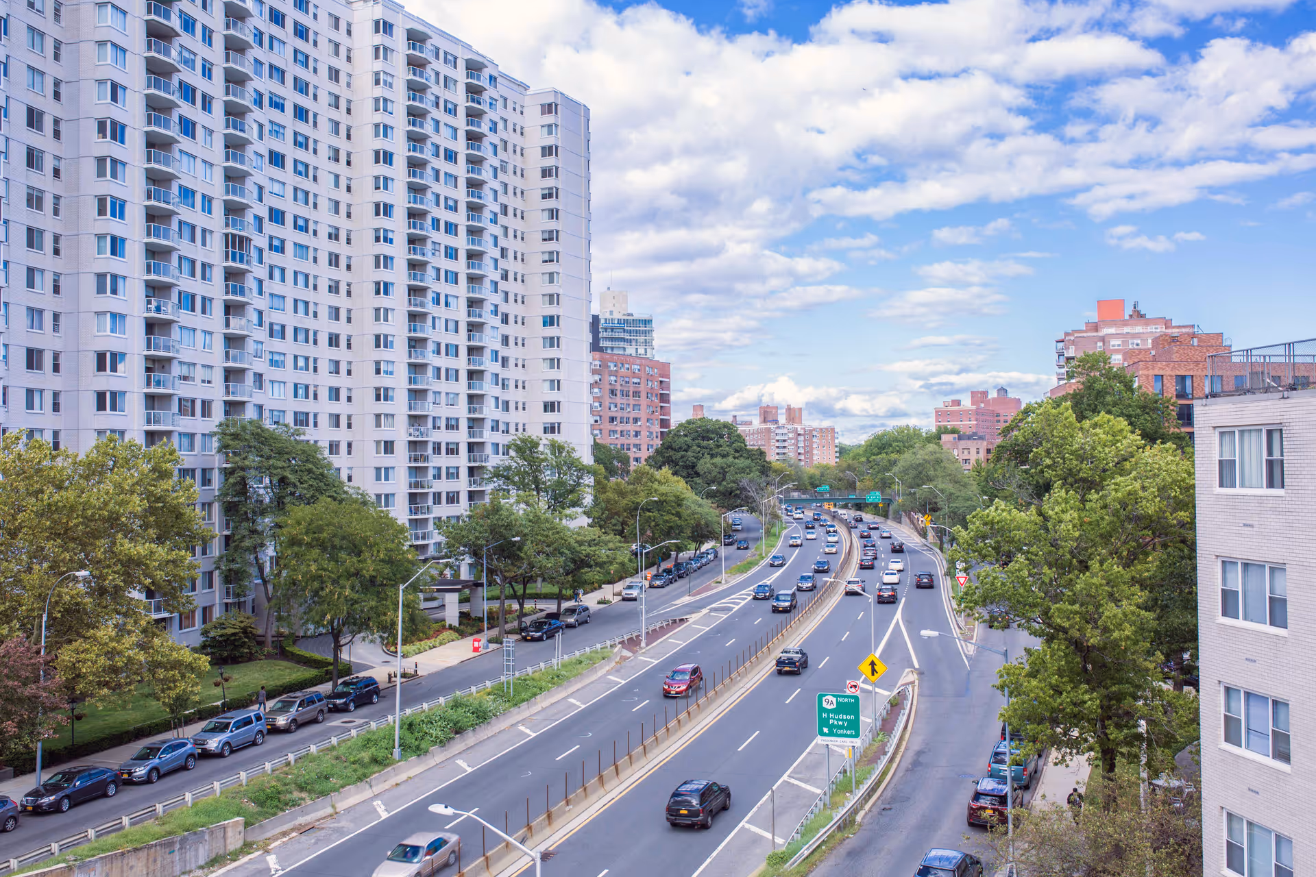 High-rise apartment buildings and trees beside a multi-lane road with cars under a partly cloudy sky.