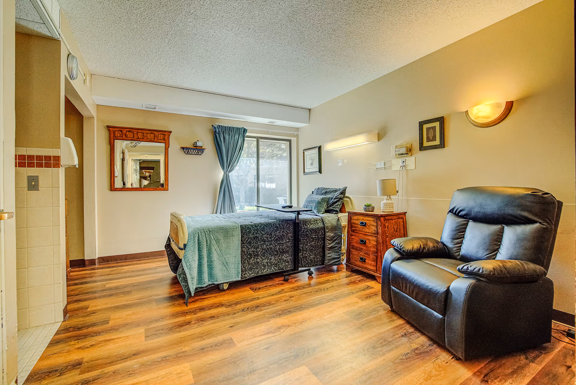 A senior living facility bedroom with a single bed covered in green and blue bedding, a wooden nightstand with a lamp, a black leather recliner chair, a wall-mounted light fixture, framed artwork on the walls, a large window with blue curtains, and wood flooring.