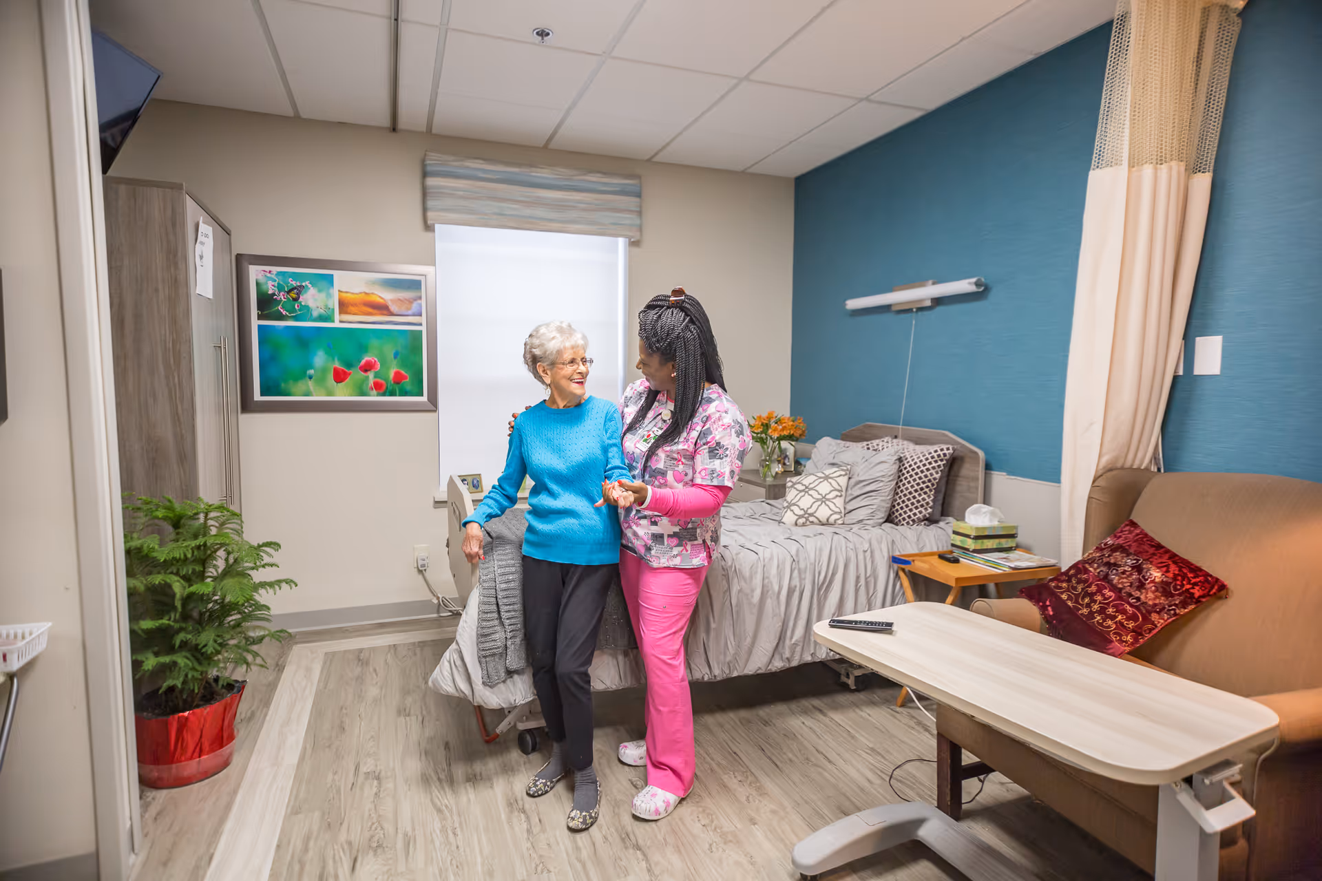An elderly woman in a blue sweater is standing and smiling while being assisted by a caregiver in pink scrubs inside a well-lit bedroom with a bed, a small table, a couch with a red pillow, and a plant in the corner.