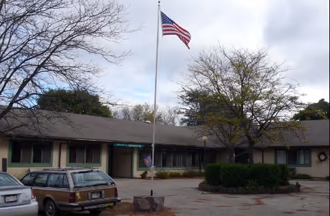 Single-story building with a gray roof and green-trimmed windows, an American flag on a flagpole in front, a tree with sparse leaves, and two parked cars in the driveway under a cloudy sky.