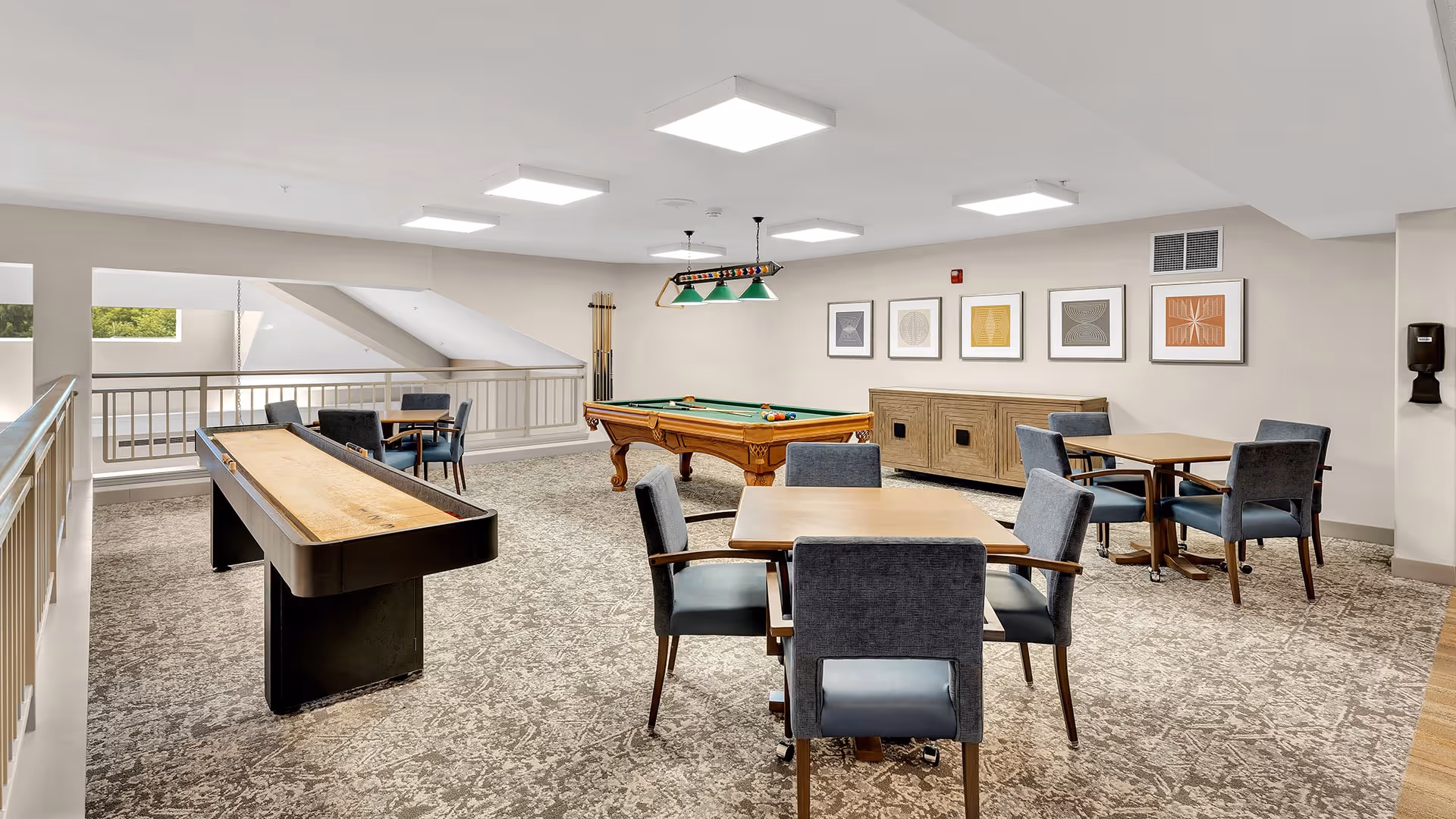 A bright and spacious recreational room with a pool table, a shuffleboard table, and several tables with chairs arranged for games or socializing. The room has light-colored walls, carpeted flooring, framed abstract artwork on the wall, and ceiling lights providing ample illumination.