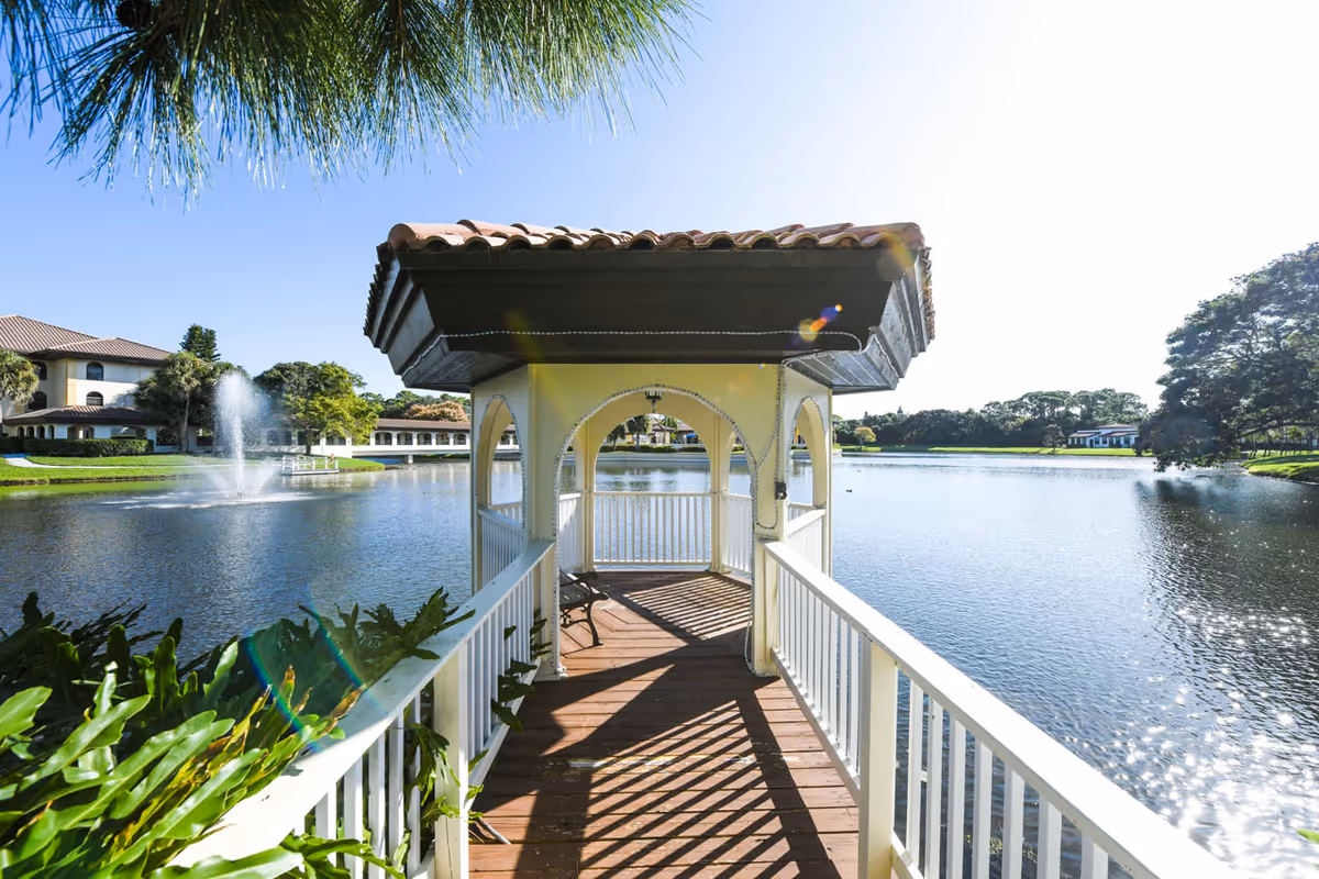 A wooden gazebo with a tiled roof extending over a lake, surrounded by greenery and buildings in the background under a clear blue sky.